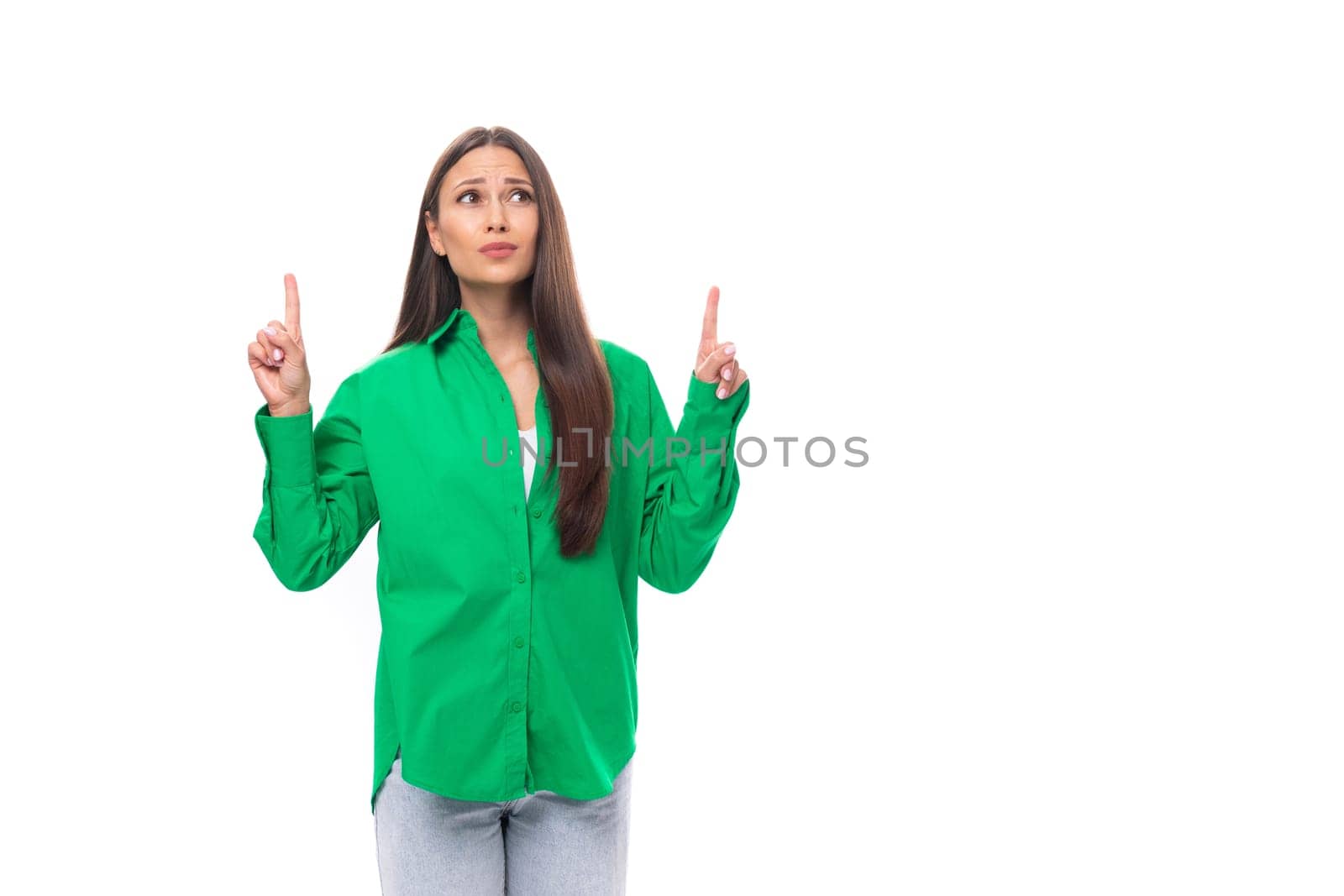 young caucasian brunette lady with make-up dressed in an elegant green shirt points with her hands towards the wall with empty space.