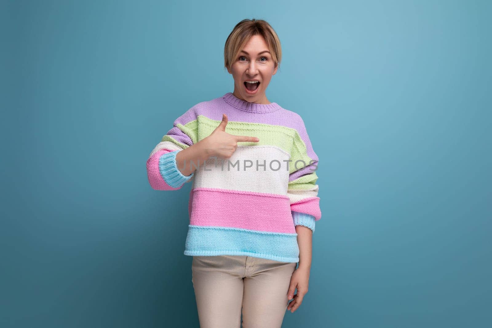 surprised blond young woman in casual outfit pointing at wall with finger on blue background with copy space.