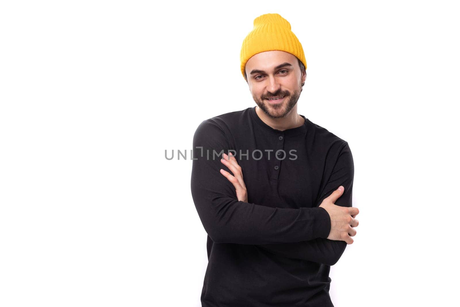 attractive young 30 year old guy with a beard dressed in a black jacket and a yellow hat looks at the camera.