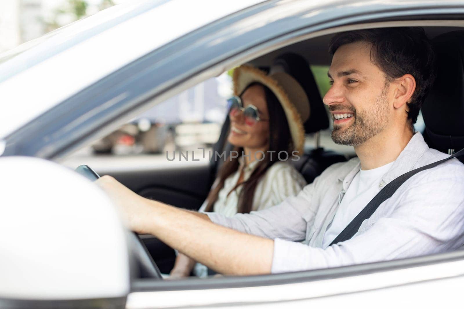 Smiling positive couple husband and wife going summer vacation by car white together. Happy millennial man and woman wearing wicker hat and sunglasses sitting in auto, enjoying road trip