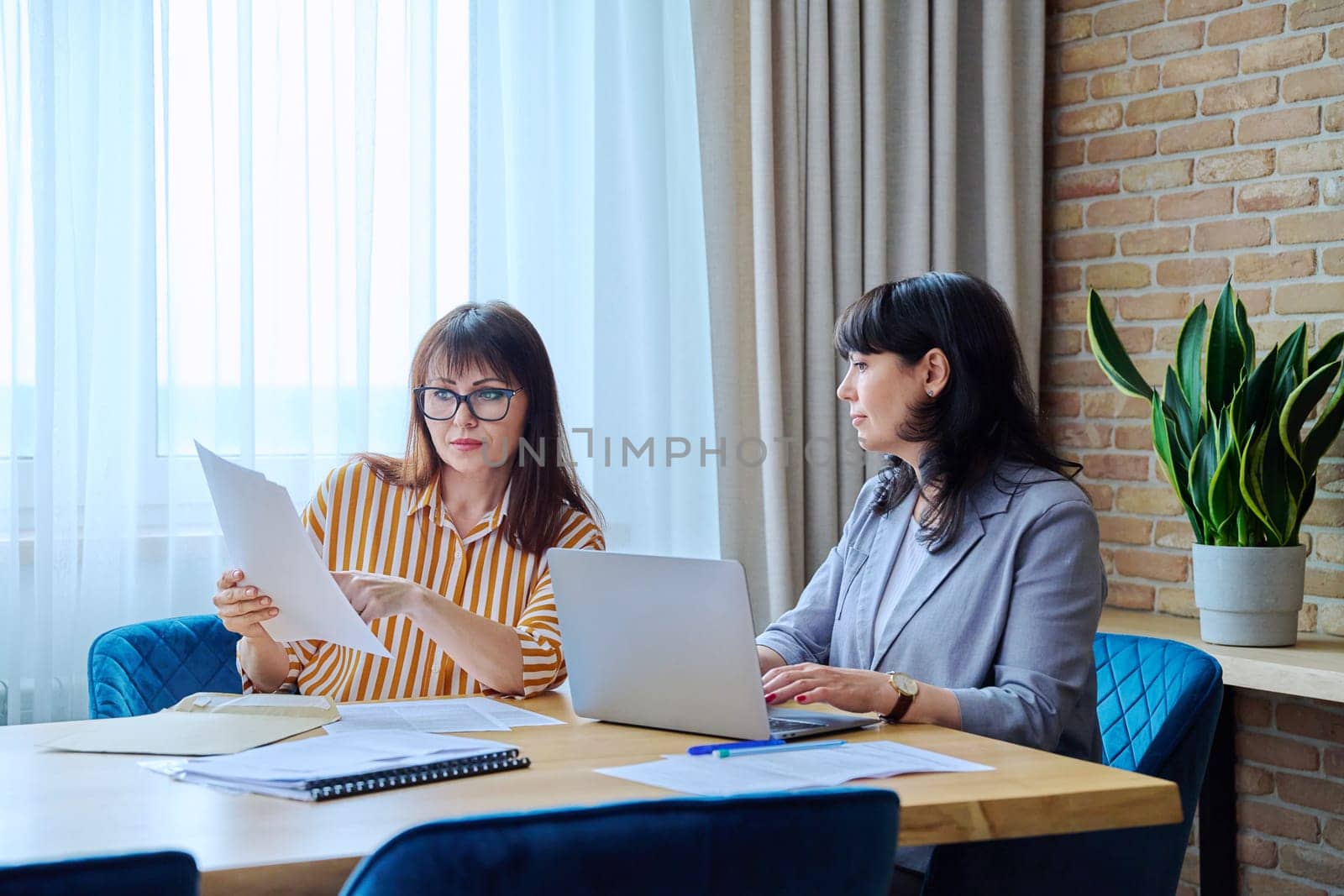 Two mature business women colleagues sitting at large table in office with business papers contracts laptop computer. Business ceo work law finance mentoring consulting teamwork, 40s 50s people on job