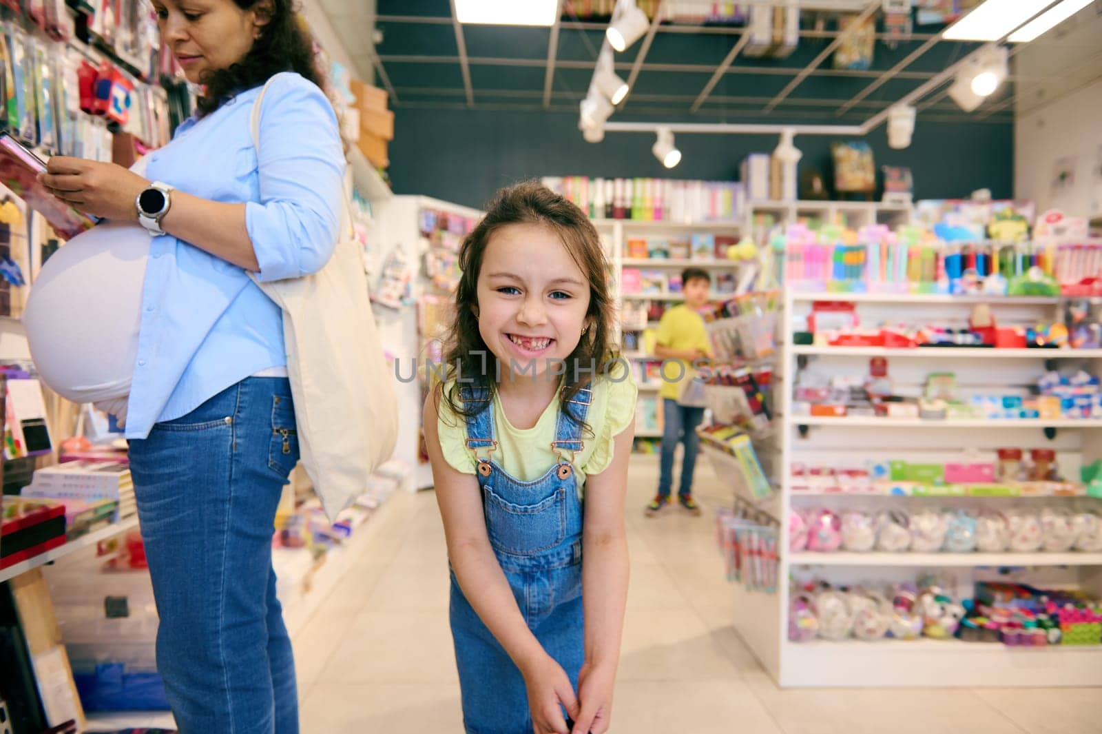 Caucasian adorable mischievous little child girl in casual denim, smiling broadly looking at camera, enjoying a shopping day with her mother in the school stationery shop, buying school supplies.