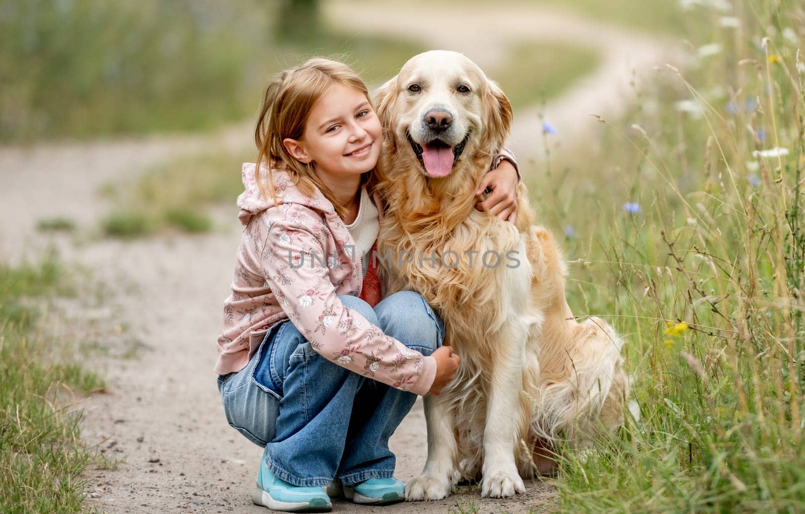 Preteen girl with golden retriever dog sitting at nature and looking back. Cute child kid hugging purebred pet doggy in park at summer