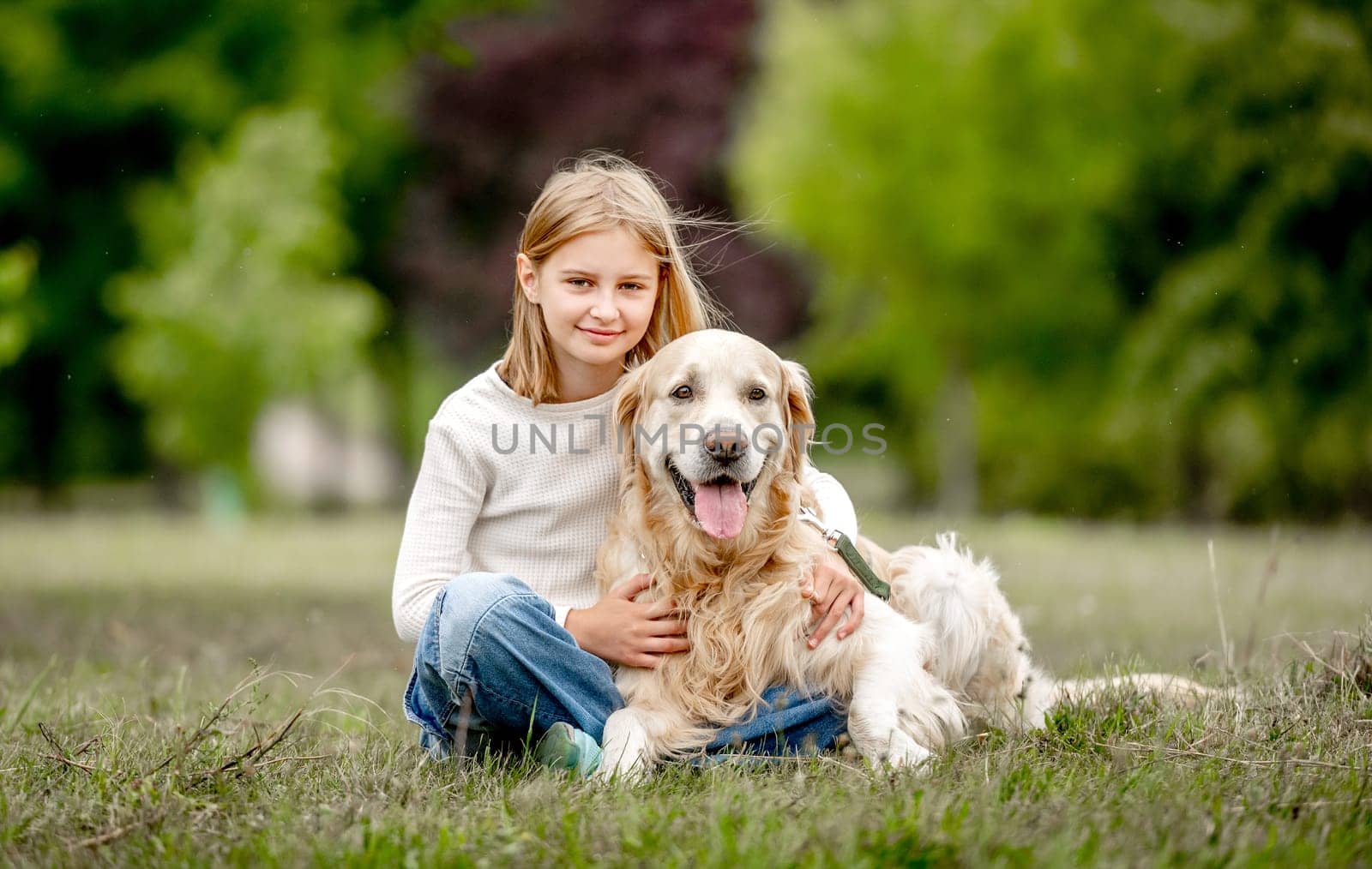 Preteen girl with golden retriever dog sitting at nature and looking back. Cute child kid hugging purebred pet doggy in park at summer
