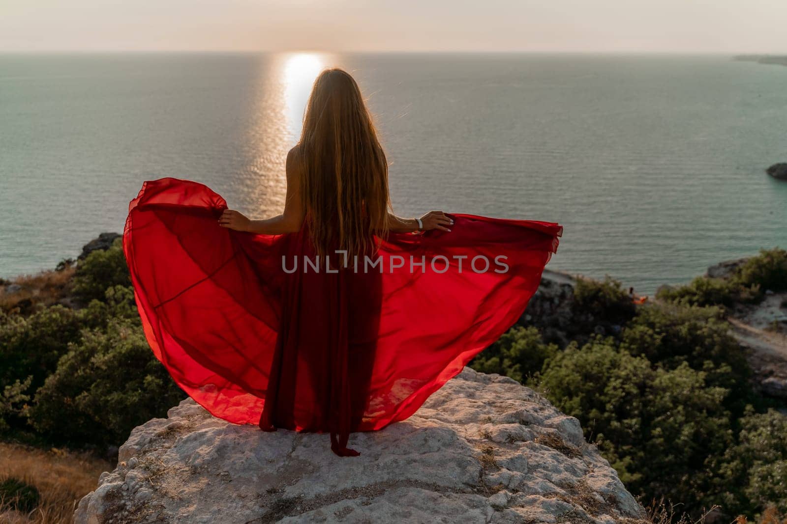 Woman sunset sea red dress, side view a happy beautiful sensual woman in a red long dress posing on a rock high above the sea on sunset