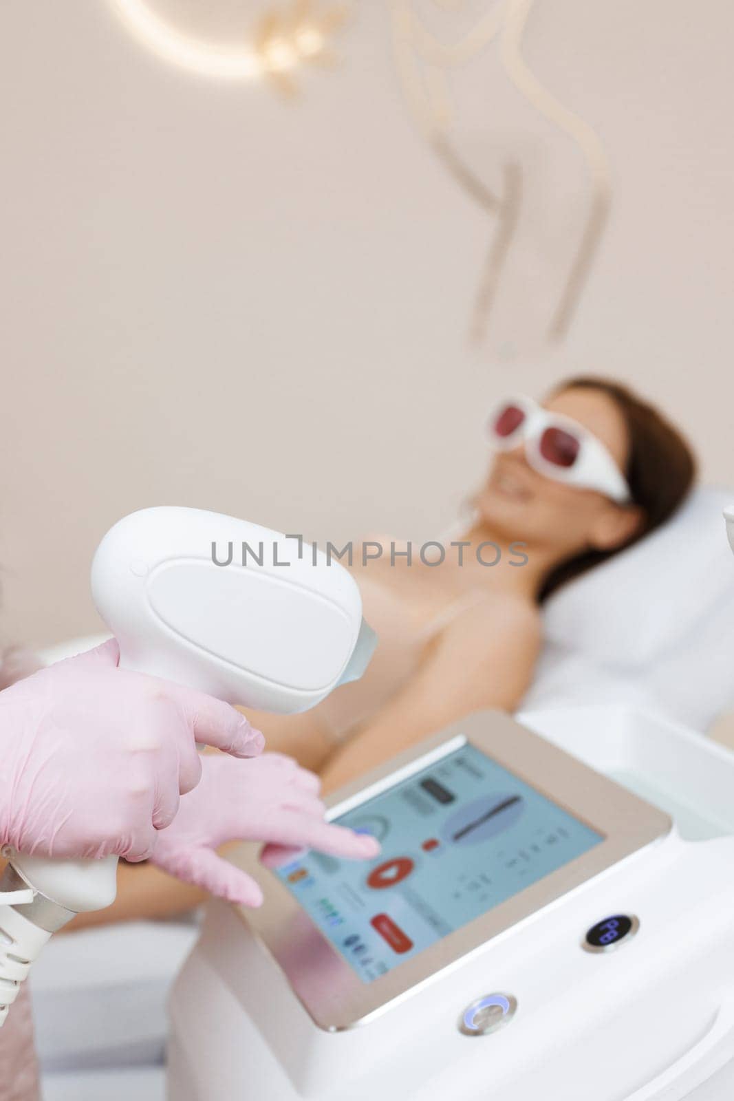 Close up of female beautician in sterile gloves using diode laser hair removal machine while smiling woman laying on daybed. Esthetician preparing equipment for epilation procedure.