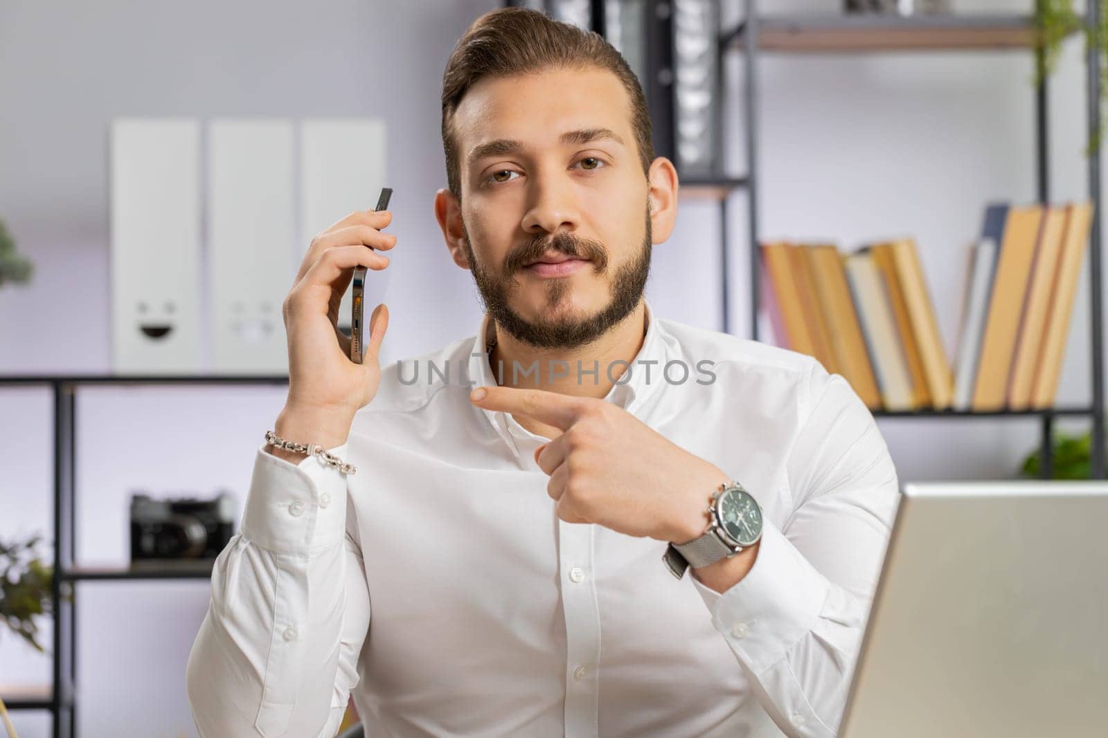 Call me. Young middle eastern boss business man showing smartphone asking to make telephone conversation, gesturing call me sign, online date. Freelancer guy at office workplace looking at camera