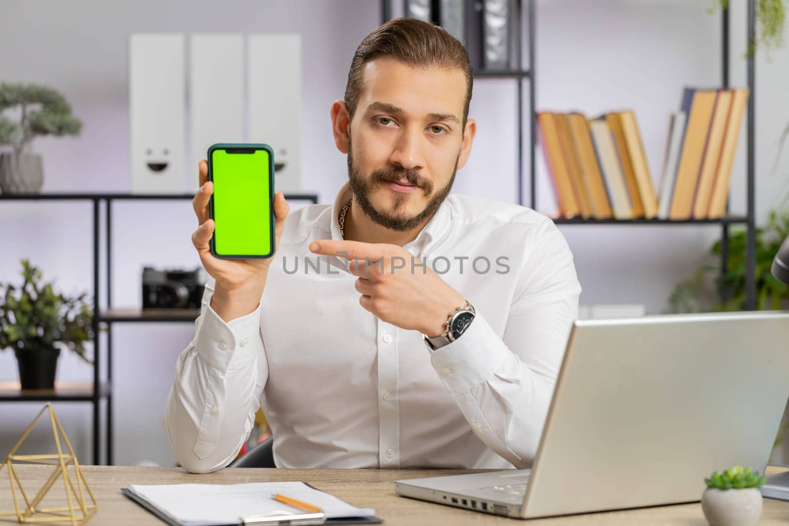 Middle eastern developer business man holding pointing on smartphone with green screen chroma key mock up recommend good application promotional sale offer. Adult freelancer guy at office workplace