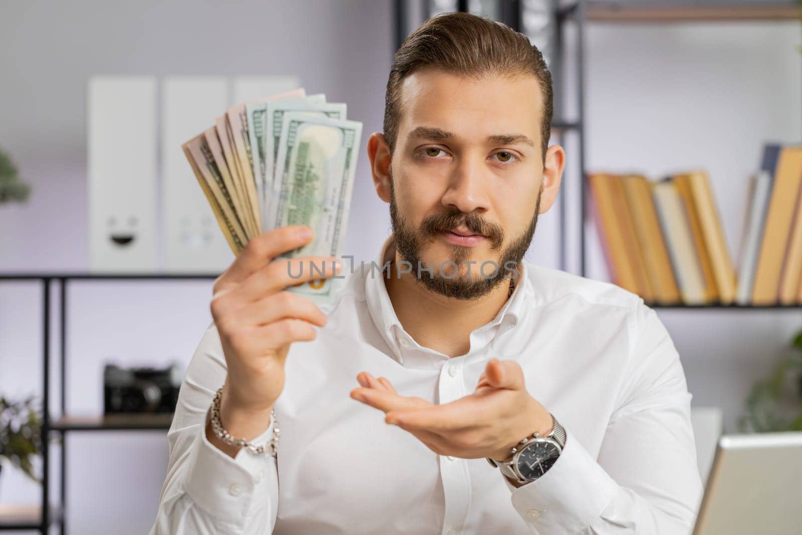Young middle eastern boss business man showing money dollar cash gives to you wages salary, satisfied with good work. Success, wealth, investment. Freelancer guy at office workplace looking at camera