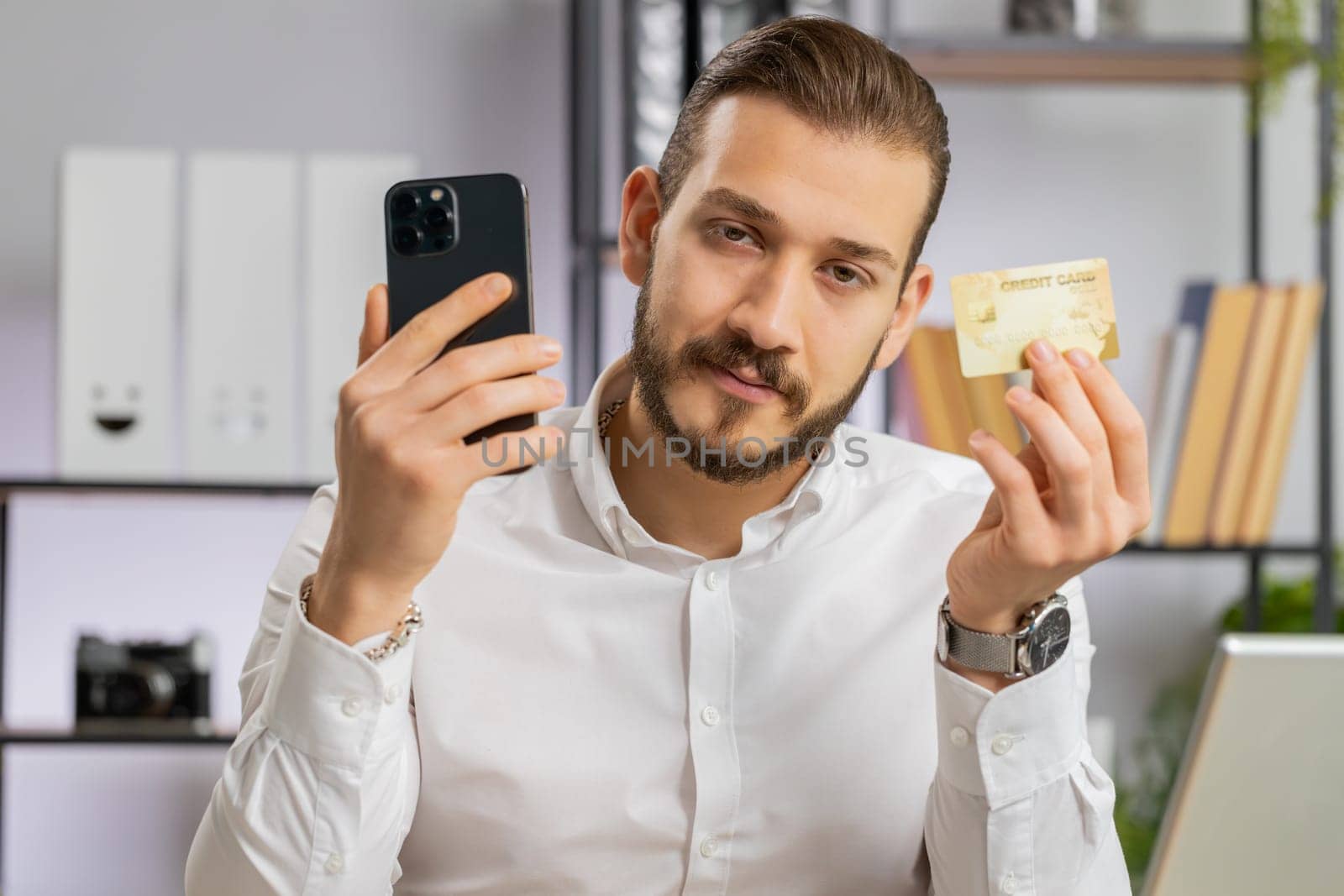 Young middle eastern boss business man using credit bank card and smartphone while transferring money, purchases online shopping ordering delivery. Freelancer guy at office workplace looking at camera