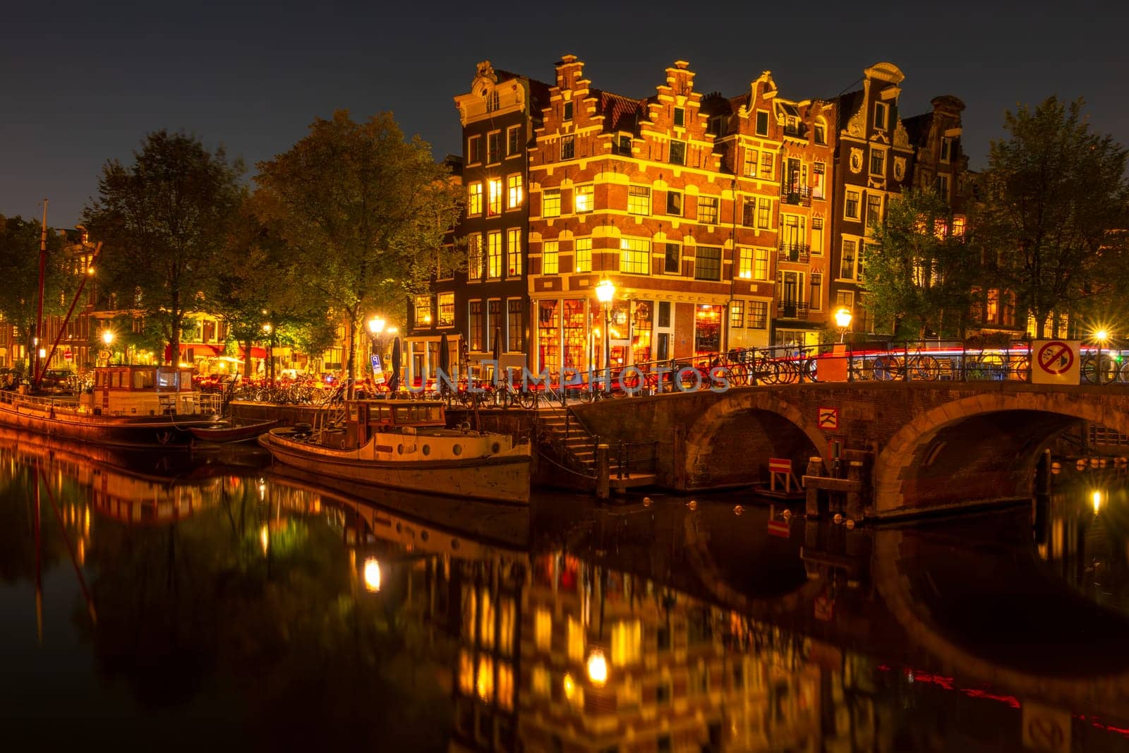 Netherlands. Summer night on the Amsterdam canal. Dancing Dutch houses with lights. Bar with outdoor tables. Moored old boats