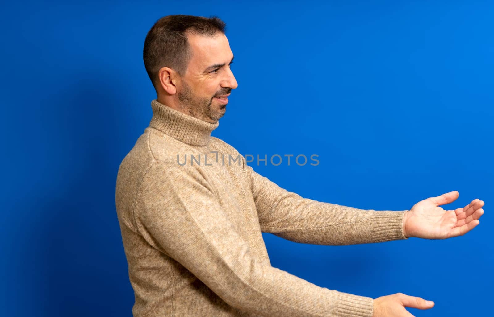 Free hugs, come into my arms. Side view of happy bearded man in his 40s stretching hands and smiling broadly, embracing, sharing love. Indoor studio shot isolated on blue background