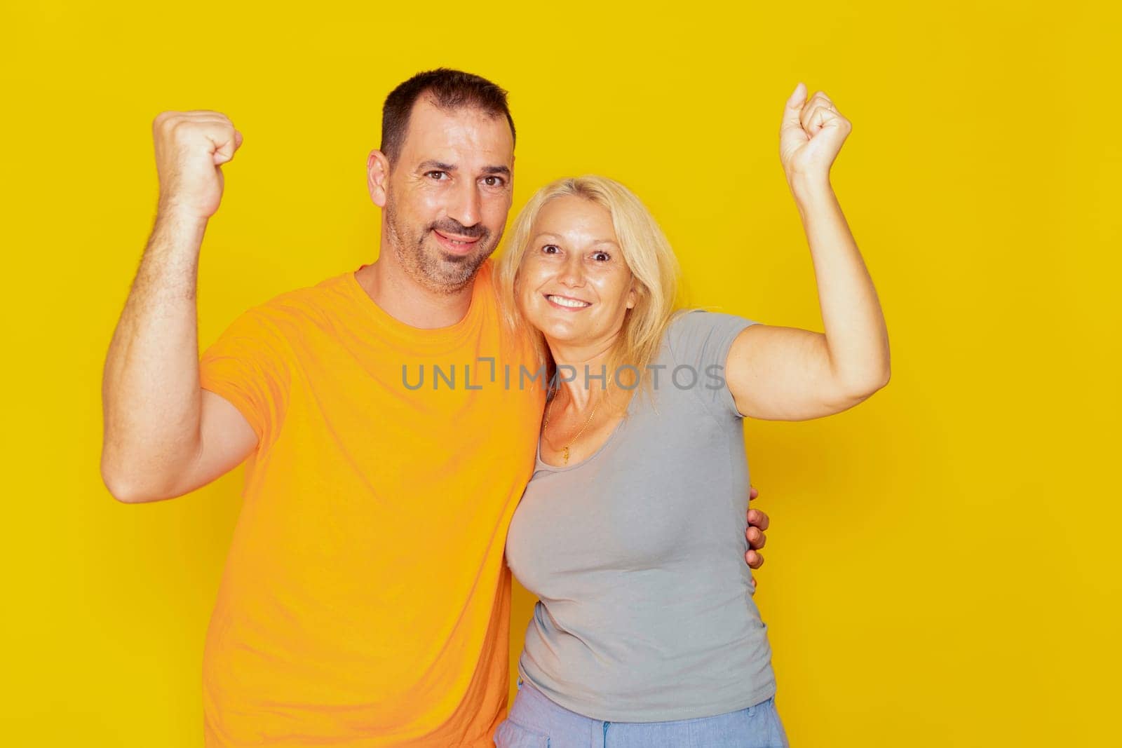 Real caucasian couple in their 40s with raised fists celebrating successful action isolated over yellow background. by Barriolo82