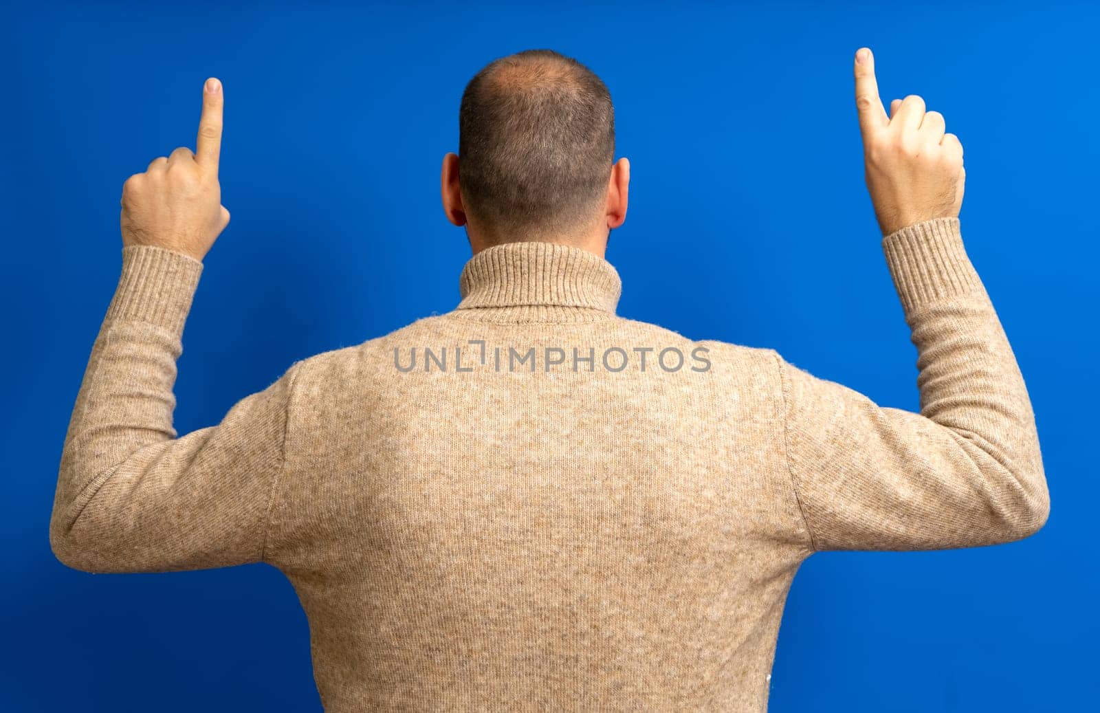 Portrait of a young man from behind, raising both hands and pointing the index finger up, wearing a beige pullover, isolated on a blue background.