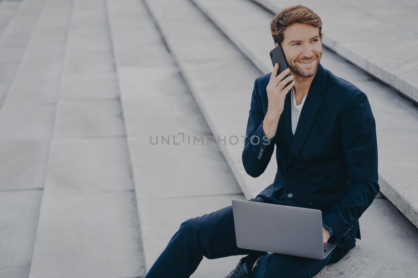 Caucasian satisfied man in formal outfit sitting on steps outdoor, talking on mobile phone and working on laptop computer. Male freelancer typing on keyboard while speaking on cellphone with client