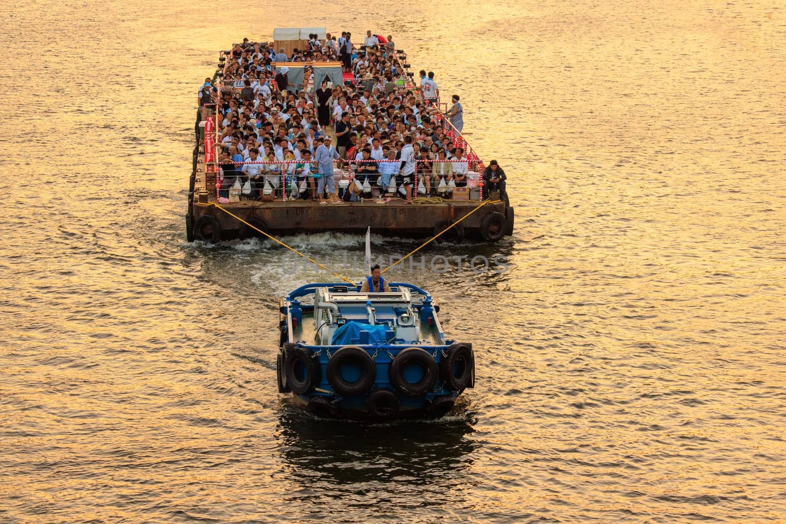 Osaka, Japan - July 25, 2023: Tug boat pulls crowded barge down river with golden glow on water by Osaze