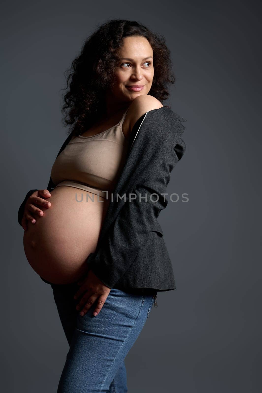 Stylish elegant pregnant woman poses bare belly, smiles looking aside, isolated on gray studio background. Happy carefree pregnancy and maternity concept. Portrait of a gravid expectant young mother