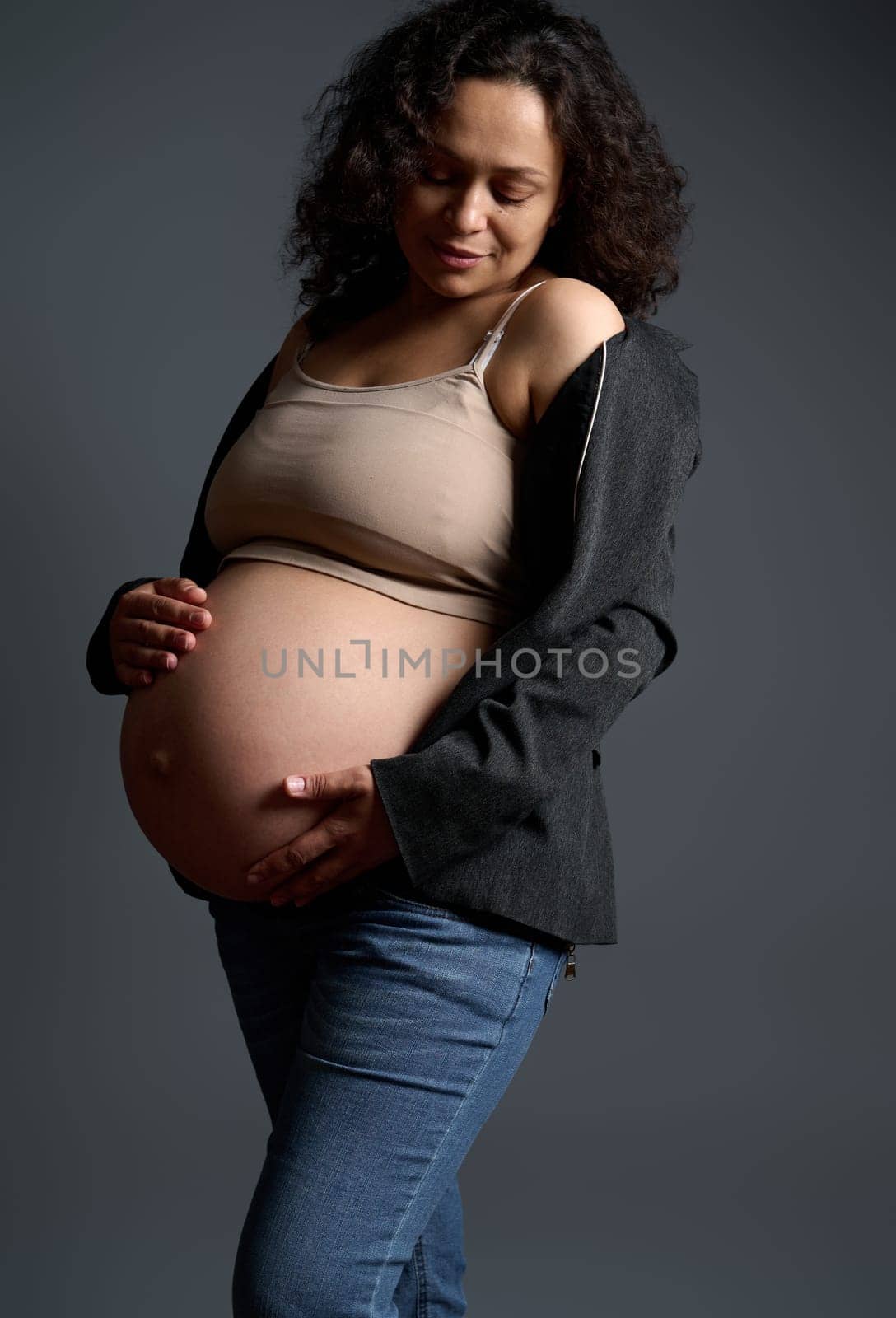 Portrait of a gorgeous pregnant woman expecting baby, putting hands on her big belly in the ninth month of happy pregnancy, experiencing positive emotions feeling baby moves, isolated gray background