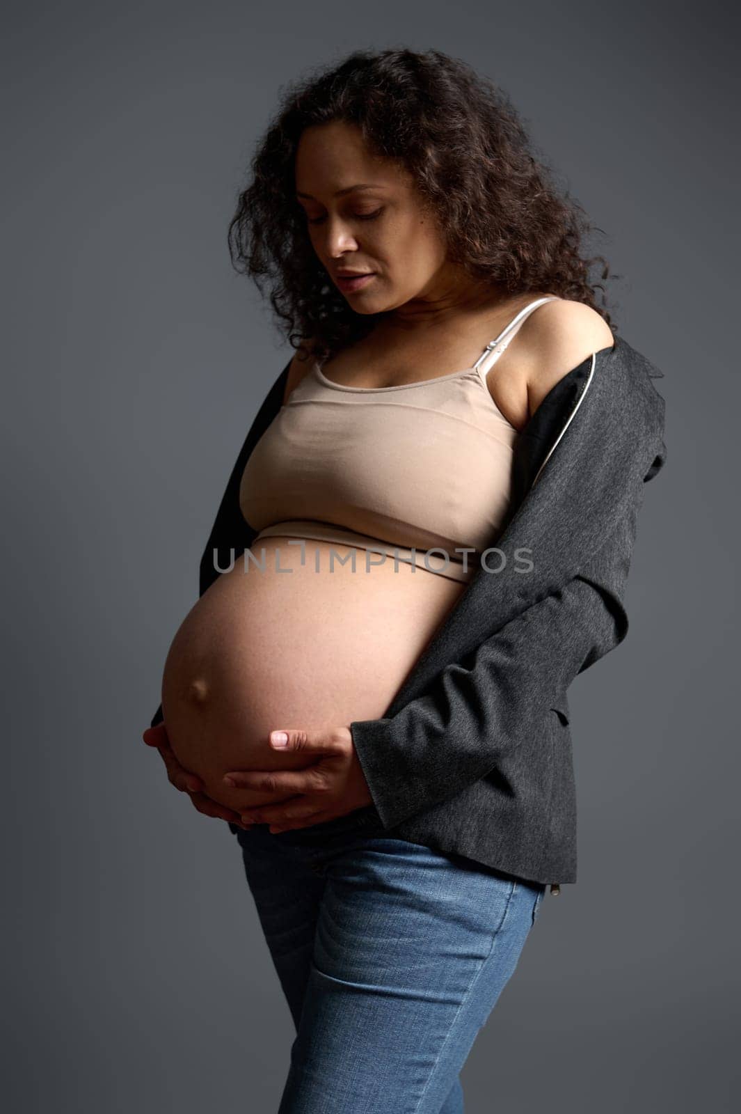 Attractive pregnant woman gently stroking her big belly, experiencing wonderful emotions feeling baby moves, isolated gray background. Happy carefree pregnancy. Maternity. Body positivity. Femininity