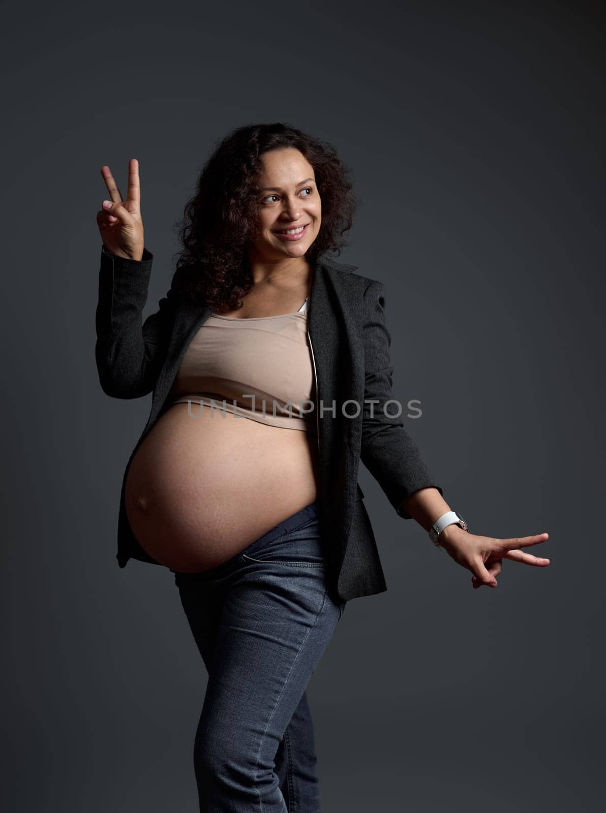 Cheerful smiling Latin American young pregnant woman, happy gravid mother shows peace sign, poses bare belly over gray isolated studio background. Pregnancy in ninth month. Maternity. Expecting a baby
