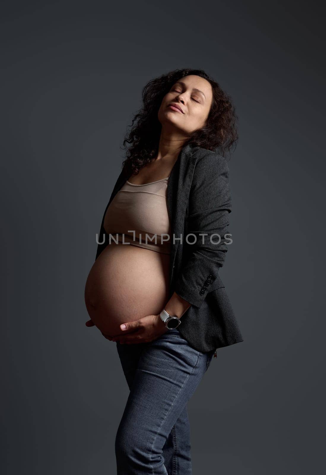 Studio portrait of a multi-ethnic woman 30s, delightful gorgeous pregnant expectant mother holding her belly, posing with her eyes closed over isolated gray background. Beautiful pregnancy. Maternity