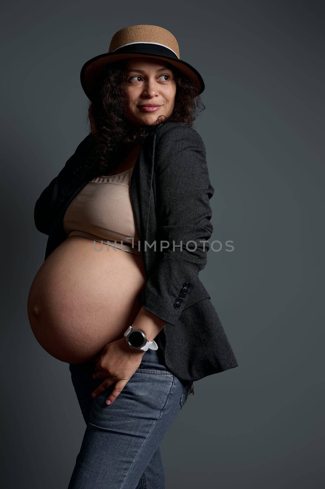 Stylish elegant pregnant woman with beautiful big naked belly, dressed in jeans, gray blazer and straw hat, smiles looking aside a copy space over studio background. The concept of beautiful pregnancy