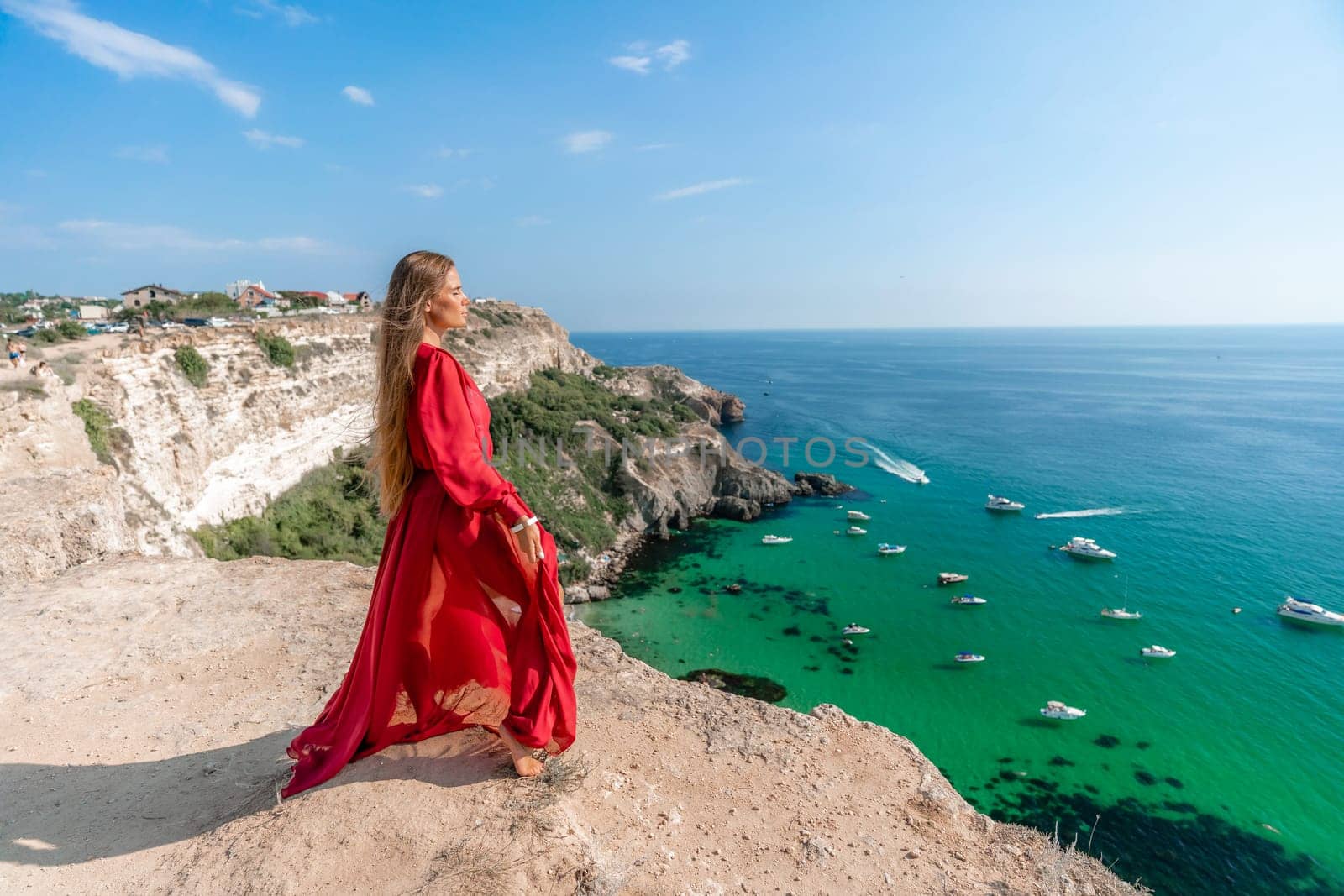 Red Dress Woman sea Cliff. A beautiful woman in a red dress and white swimsuit poses on a cliff overlooking the sea on a sunny day. Boats and yachts dot the background