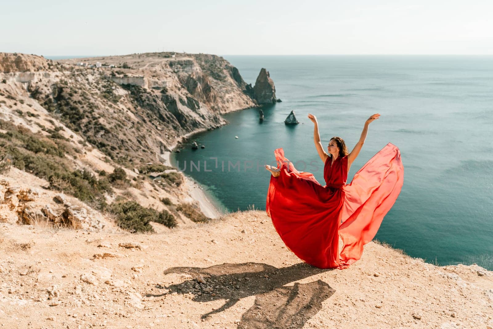 Woman red dress sea. Female dancer posing on a rocky outcrop high above the sea. Girl on the nature on blue sky background. Fashion photo