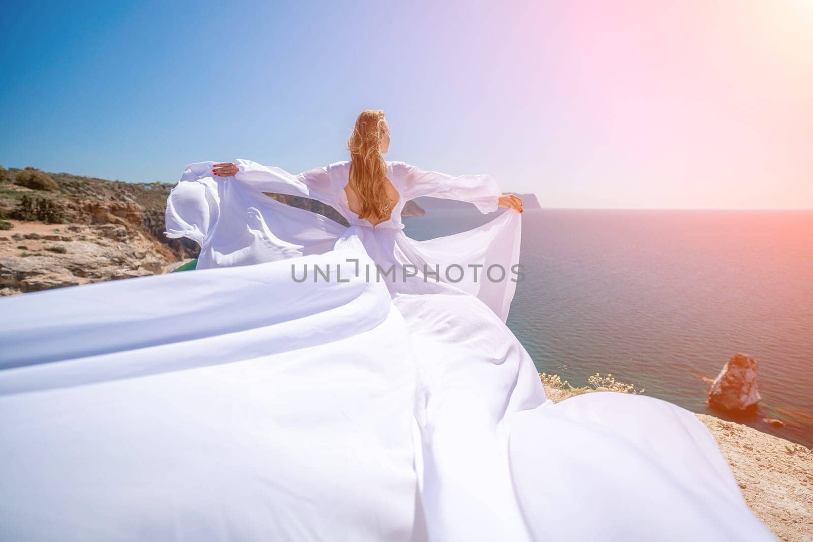 woman sea white dress. Blonde with long hair on a sunny seashore in a white flowing dress, rear view, silk fabric waving in the wind. Against the backdrop of the blue sky and mountains on the seashore