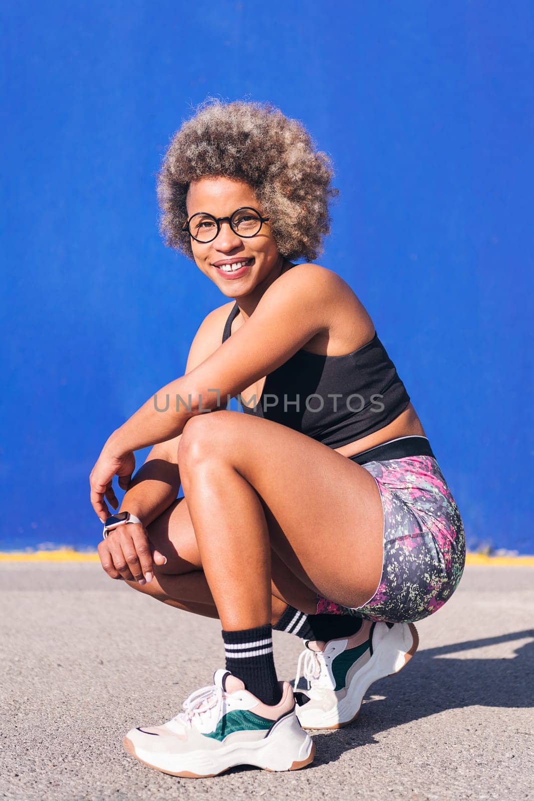 vertical portrait of an african american sportswoman squatting looking at camera smiling happy in a blue background, concept of sport and healthy lifestyle