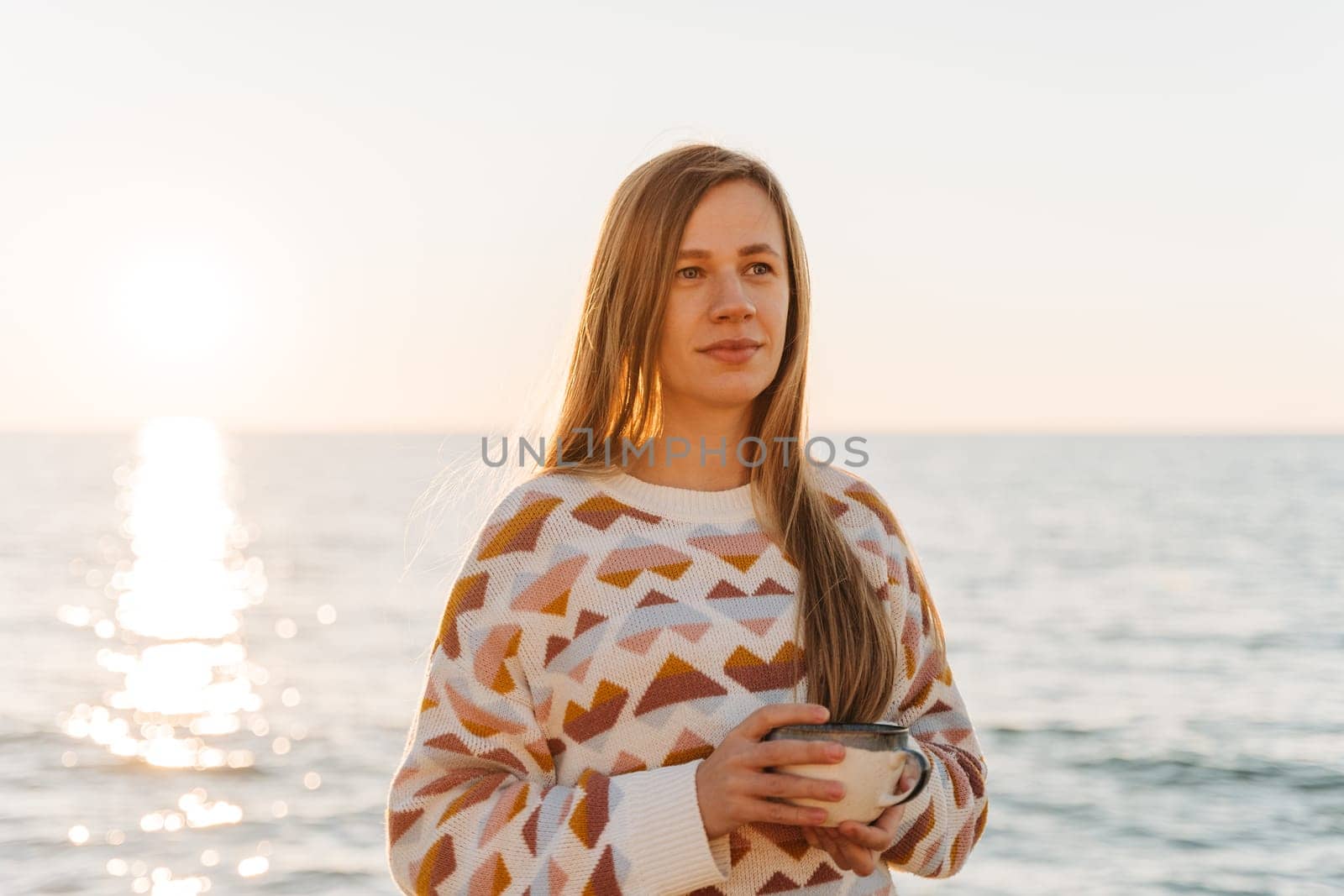 Young beautiful girl in cozy sweater holding coffee cup while enjoying winter sun on seaside shore during mild sunset. Cute attractive woman enjoying cup of tea with autumn ocean sun trail background.