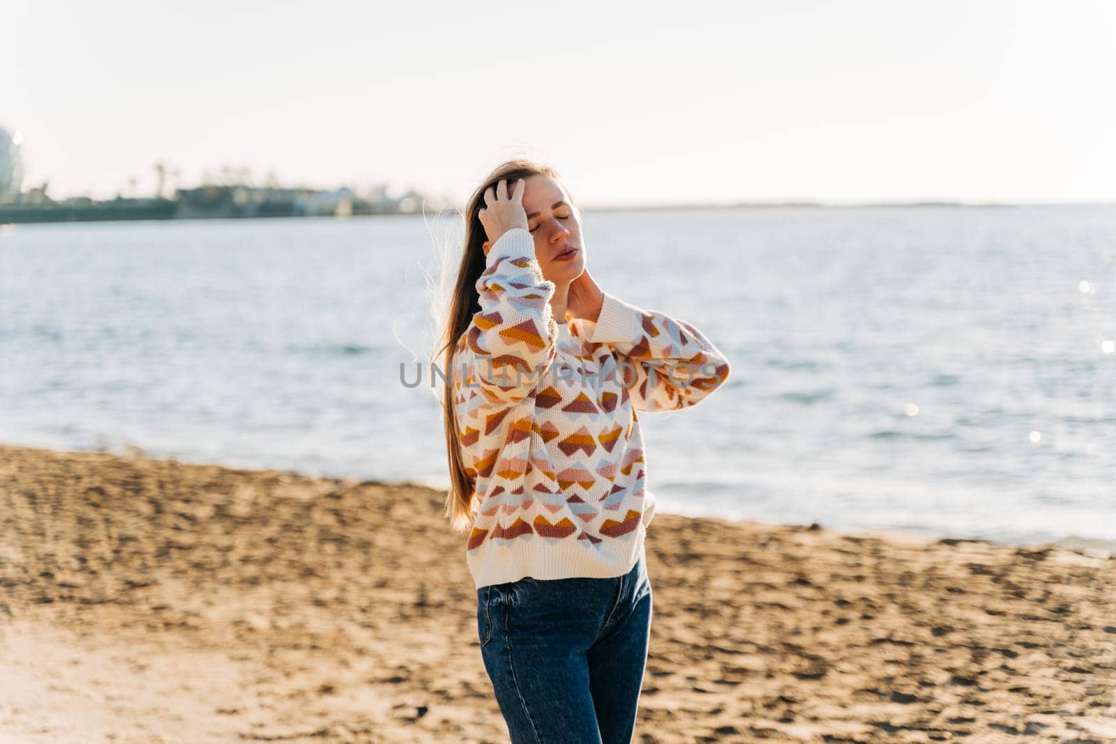 Young beautiful woman in cozy sweater smiling and enjoying sunbathe on the sand beach near winter ocean. Cute attractive girl relaxing under the sun on autumn seaside shore.