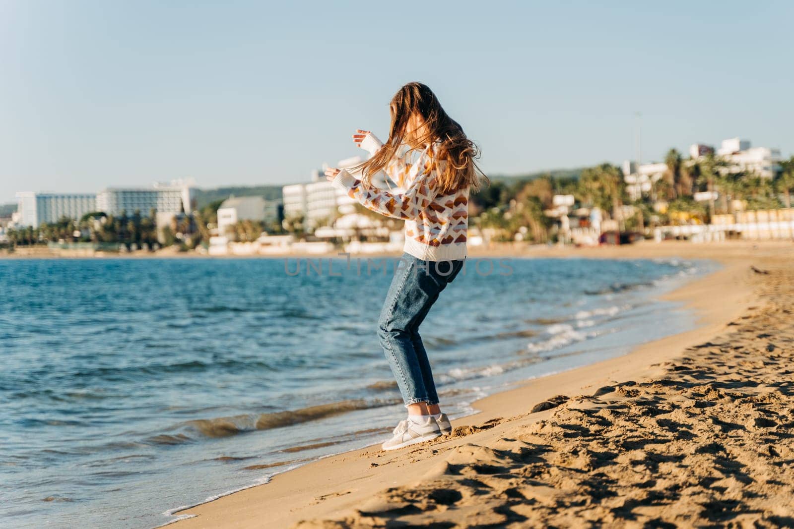 Side portrait of young beautiful woman in cozy sweater running to the winter sea waves. Cute attractive girl having fun jumping near autumn ocean on the beach sand.