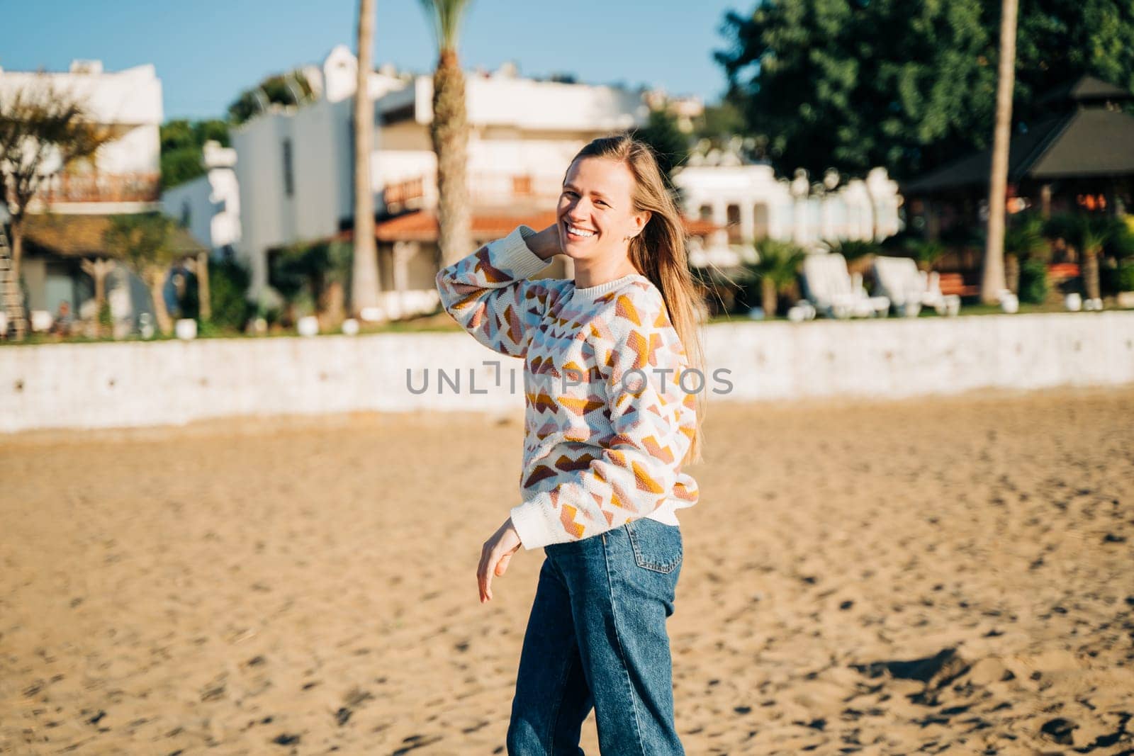 Young beautiful woman in cozy sweater smiling enjoying winter sunbathe on the sand beach with white beach bungalows in the background. Cute attractive girl relaxing under sun on autumn seaside shore by Ostanina