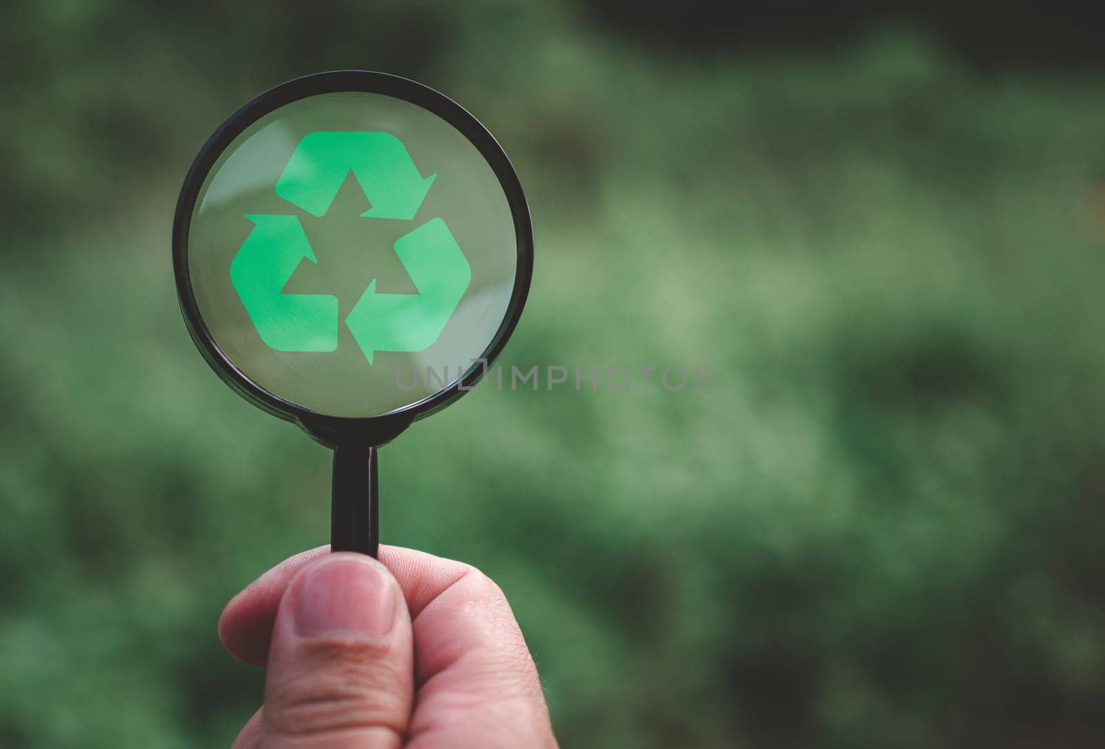 Concepts of waste reduction, pollution, reuse, efficient use of resources. Protect the environment by recycling. Human hand holding a magnifying glass with recycling sign on nature background.
