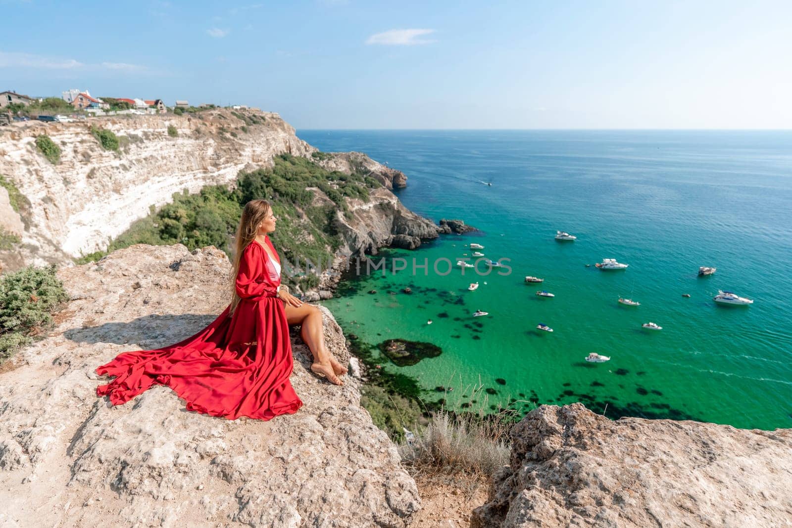 Woman red dress sea. Happy woman in a red dress and white bikini sitting on a rocky outcrop, gazing out at the sea with boats and yachts in the background