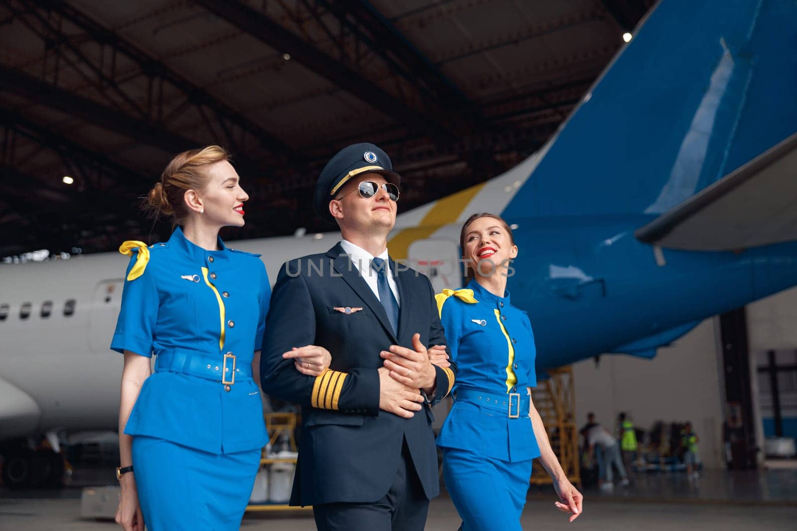 Happy pilot in uniform and aviator sunglasses walking together with two air stewardesses in blue uniform in front of big passenger airplane in airport hangar. Aircraft, occupation concept