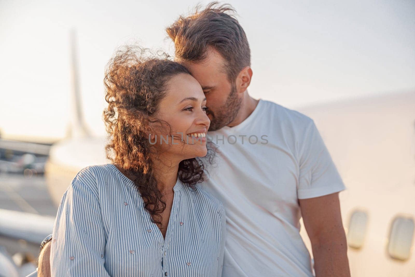 Portrait of lovely couple of tourists, man and woman looking happy while standing outdoors ready for boarding the plane at sunset. Vacation, lifestyle, traveling concept