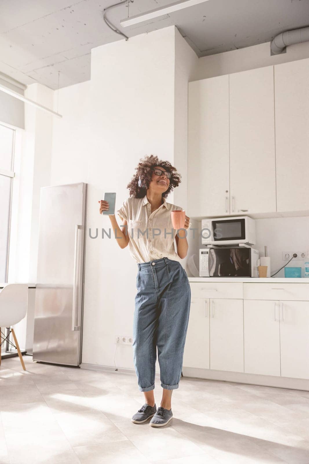 Full-length photo of cheerful female using wireless headphones and dancing on the kitchen while holding phone