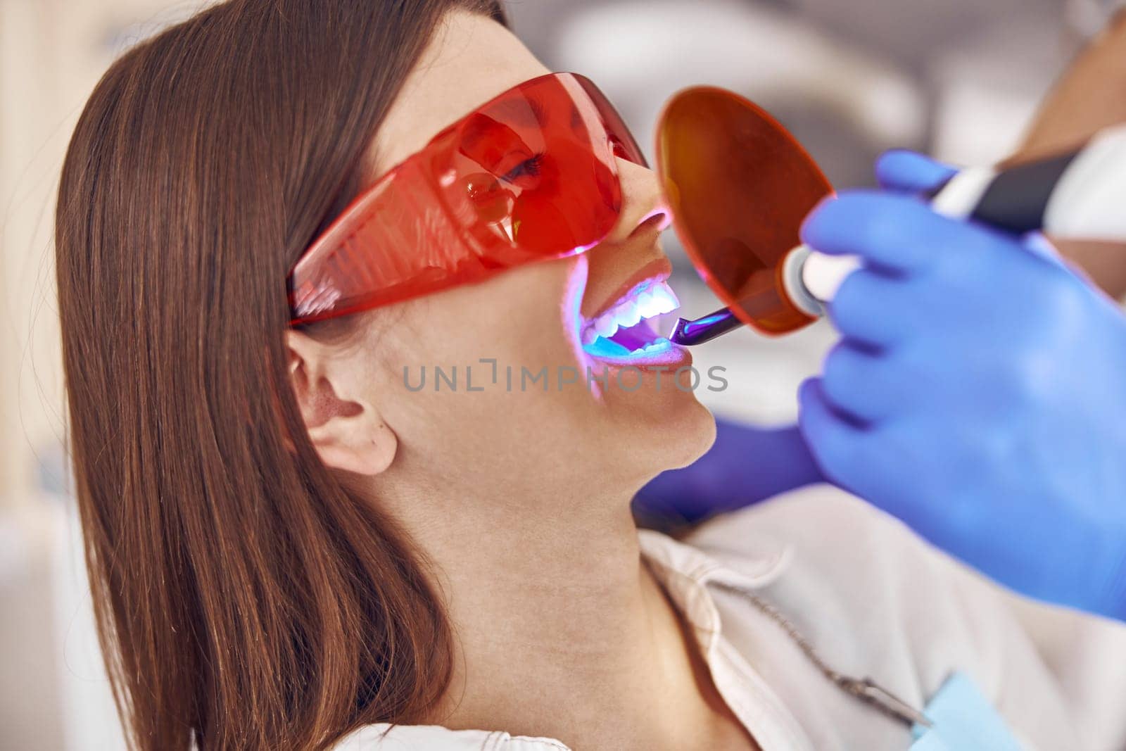 Professional male doctor uses ultraviolet lamp after making a filling on a tooth for joyful happy caucasian woman in modern dental cabinet