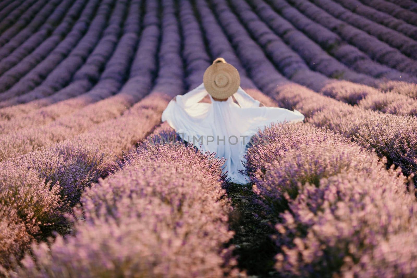 Happy woman in a white dress and straw hat strolling through a lavender field at sunrise, taking in the tranquil atmosphere. by Matiunina