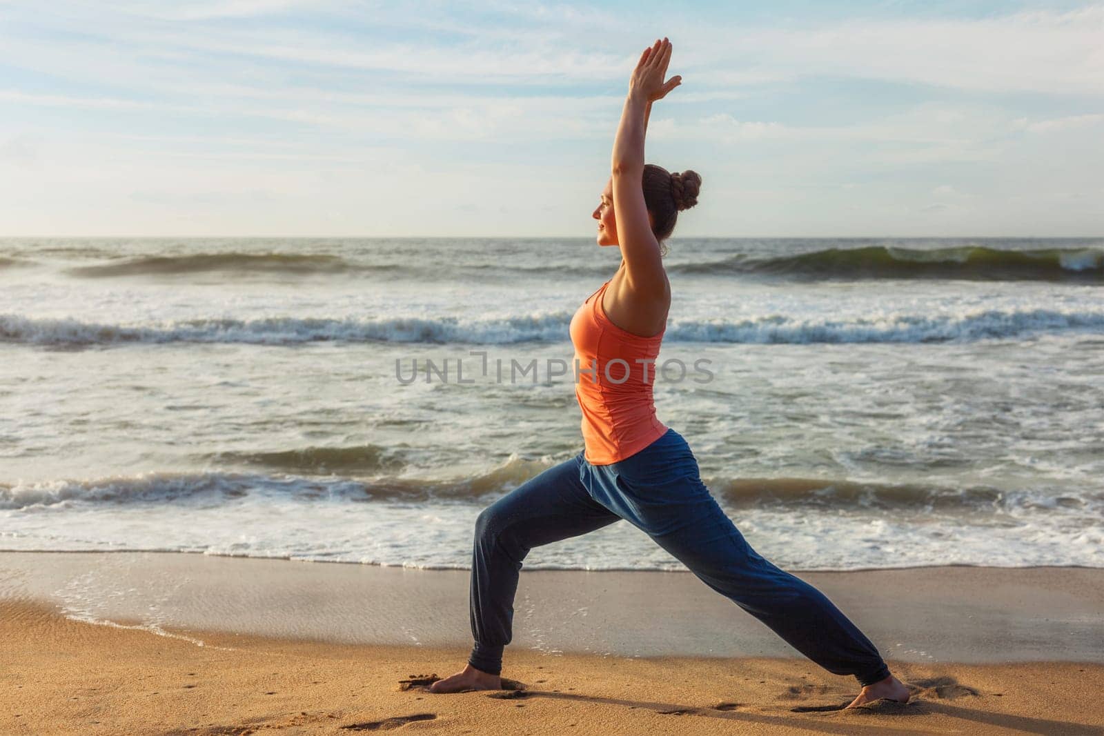 Woman doing yoga asana Virabhadrasana 1 Warrior Pose on beach on by dimol