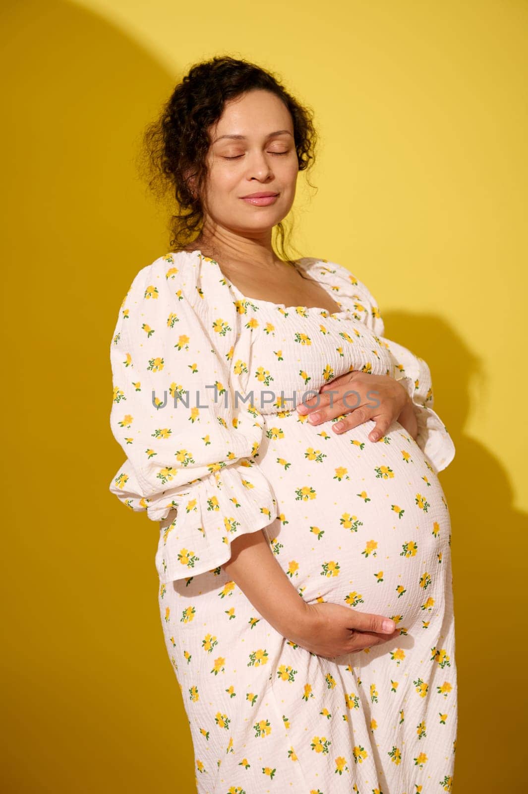 Happy pregnant woman, gravid expectant mother in stylish white summer sundress, touching her big belly, posing with her eyes closed, enjoing her pregnancy and maternity, isolated on yellow background.