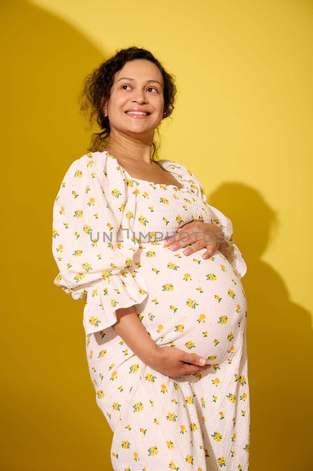 Multi-ethnic smiling middle aged pregnant woman, gravid expectant mother in stylish patterned summer sundress, touching her big belly, isolated over yellow studio background. Beautiful pregnancy
