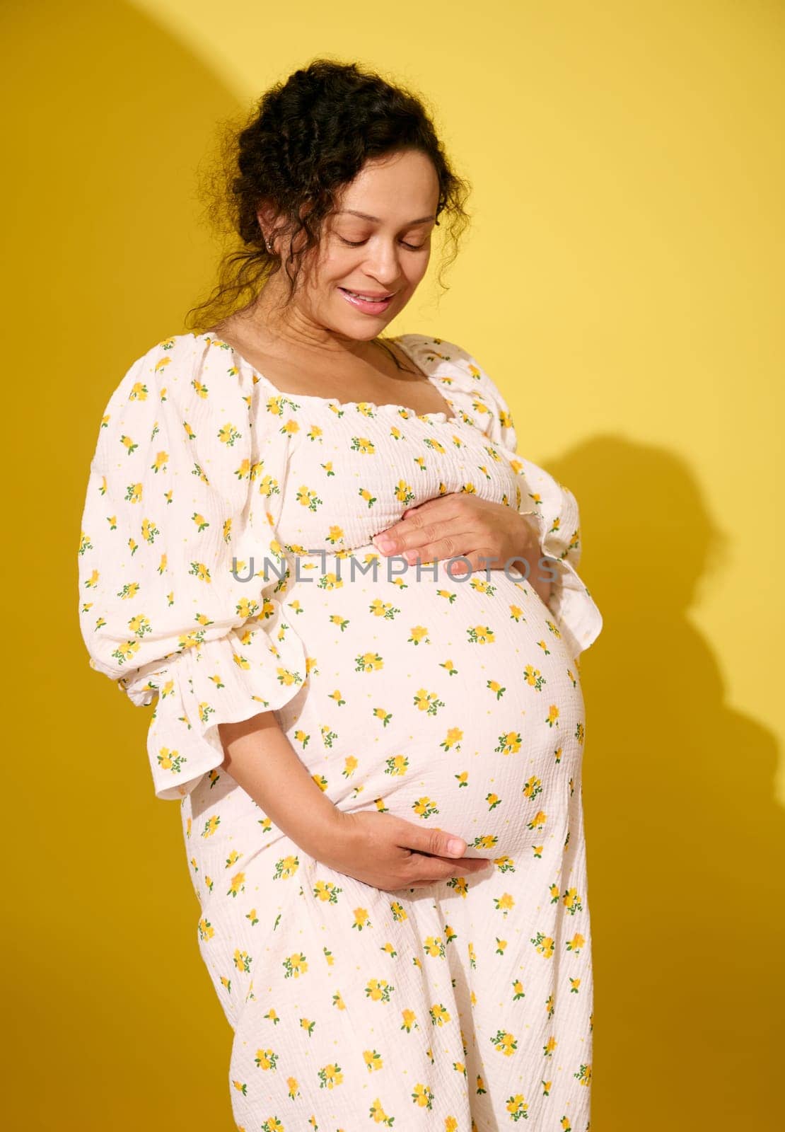 Multi-ethnic curly haired middle aged pregnant woman, gravid expectant mother in stylish patterned summer sundress, stroking her big belly, isolated over yellow studio background. Beautiful pregnancy