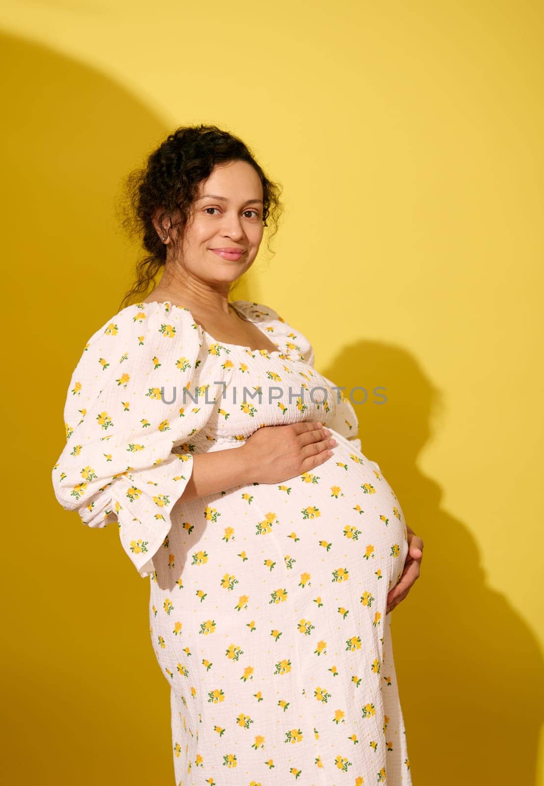 Studio portrait of a beautiful multi-ethnic pregnant curly haired woman, dressed in white summer dress, cutely smiling looking at camera, holding her belly, isolated over yellow studio background.