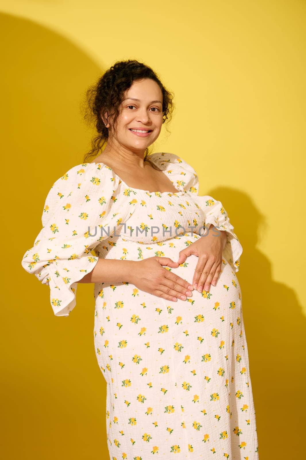 Beautiful pregnant woman, gravid expectant mother, dressed in summer sundress, holding hands on her big belly, making a shape of heart, isolated over yellow studio background. Happy carefree pregnancy
