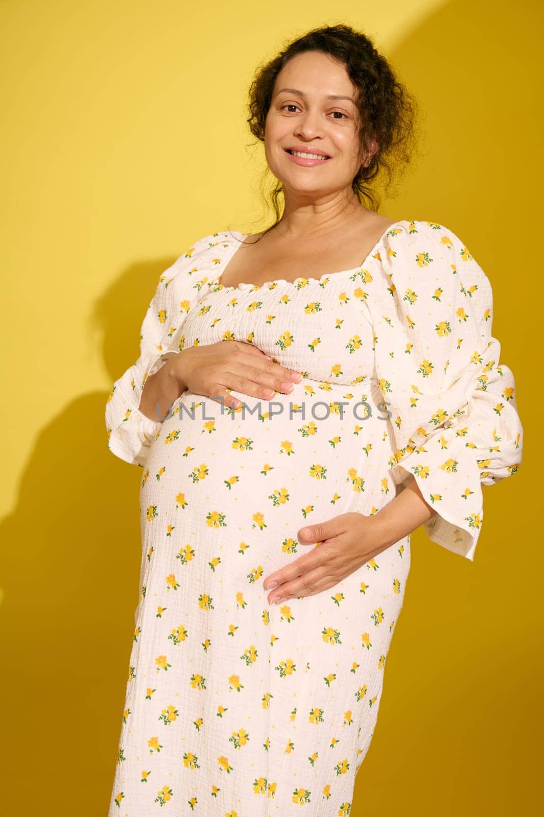 Beautiful multi-ethnic curly haired pregnant woman in stylish summer sundress, gently caresses her belly, smiles looking at camera, isolated on yellow background. Expecting a baby. Childbearing.