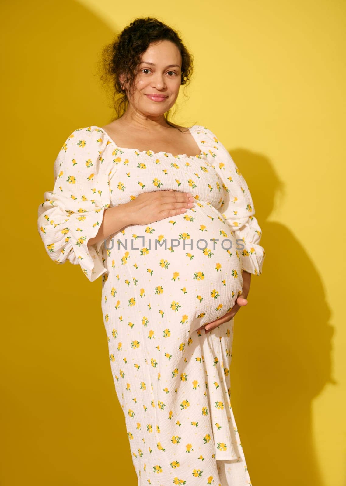 Beautiful curly haired pregnant woman in stylish summer sundress, gently caresses her belly, smiles looking at camera, isolated on yellow background. Beautiful pregnancy and maternity leave concept