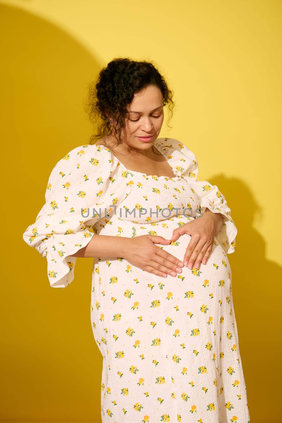 Beaautiful pregnant woman in sundress holding her hands on her big belly, making a shape of heart, isolated over yellow studio background
